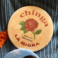 Two buttons on a textured fabric background, one with 'chinga LA MIGRA' text and a rose, the other with a heart design in red, white, and green.