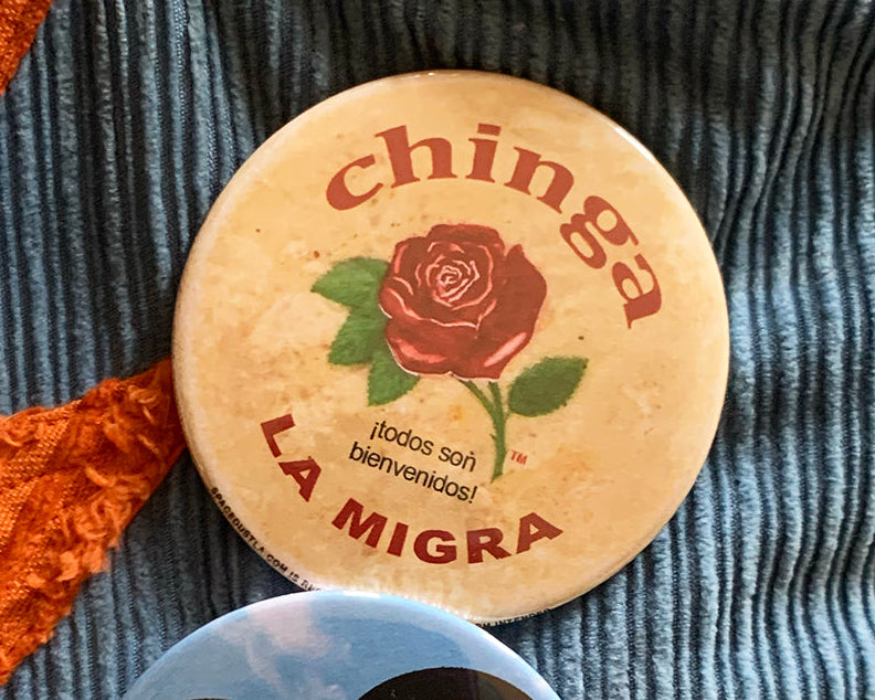 Two buttons on a textured fabric background, one with 'chinga LA MIGRA' text and a rose, the other with a heart design in red, white, and green.
